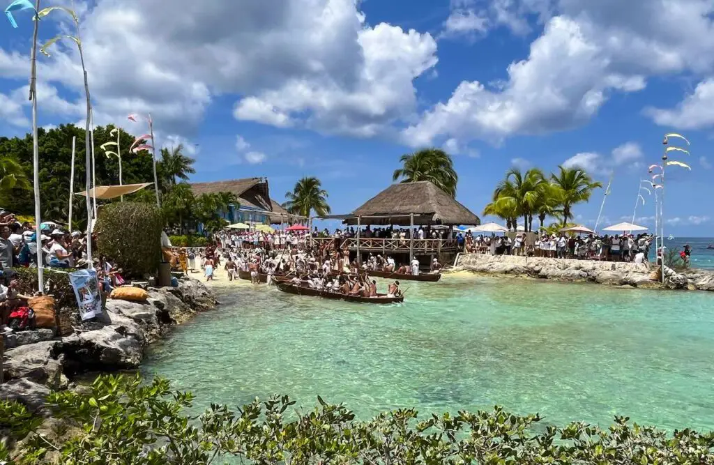 Many reenactment canoes arrive to Cozumel's Chankanaab shore with palapas, palm trees, and blue skies in the background from 2023 event.