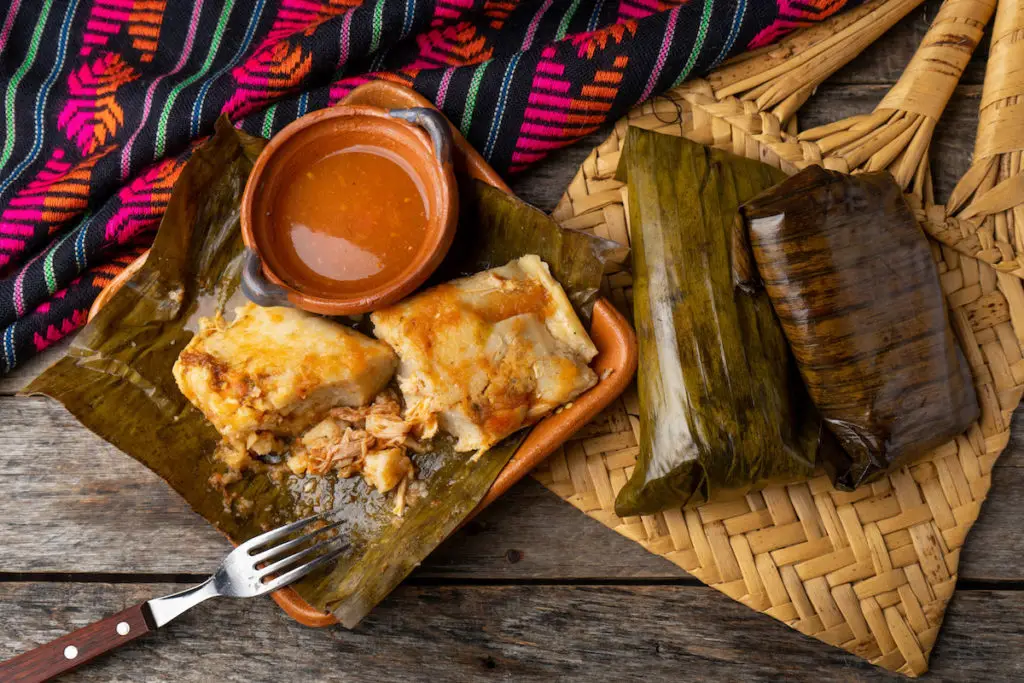 Mexican tamales shown wrapped in banana leaves, with side of hot sauce