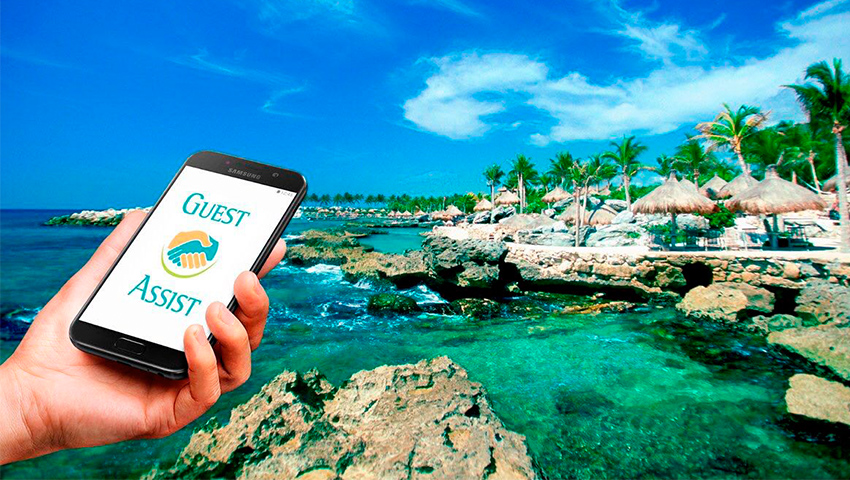 Image of a hand holding a phone with the Guest Assist app on the screen, with a backdrop of Caribbean shoreline with palapas and palm trees. 
