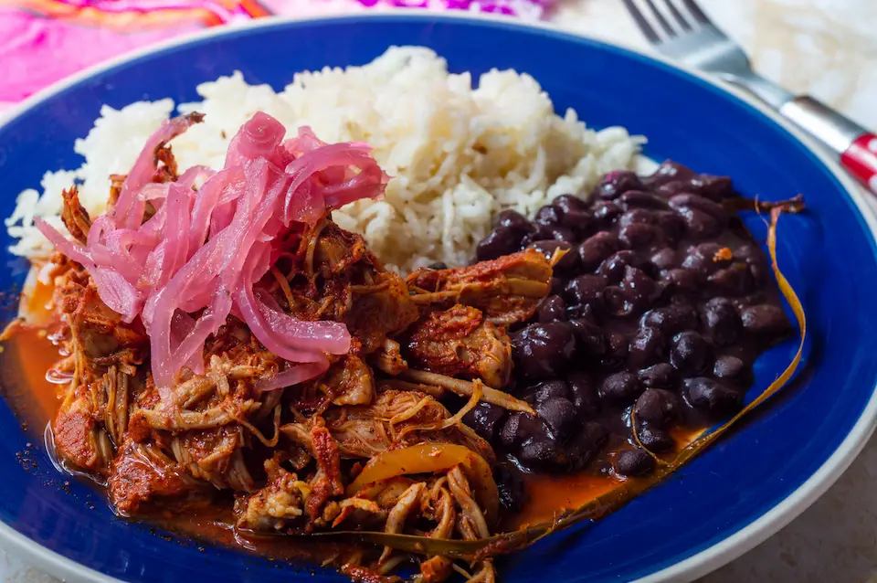 Plate of traditional cochinita pibil from the Yucatan peninsula with a side of black beans, rice, and pickled red onion. 