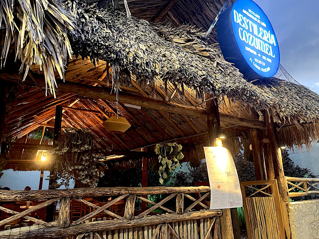 Image of La Destilería Cozumel, a new cafe with house distilled rum and gin. Photo shows blue lit sign and thatched palapa roof. 