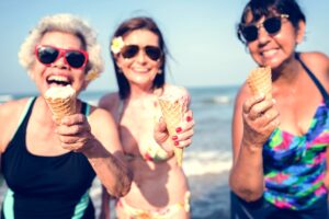 Three women smiling and laughing while eating ice cream cones at the beach.