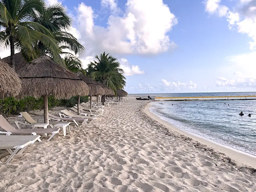 Image of Nachi Cocom beach front, showing loads of empty lounge chairs and individual palapa umbrellas near the sandy shore entry to the water. 