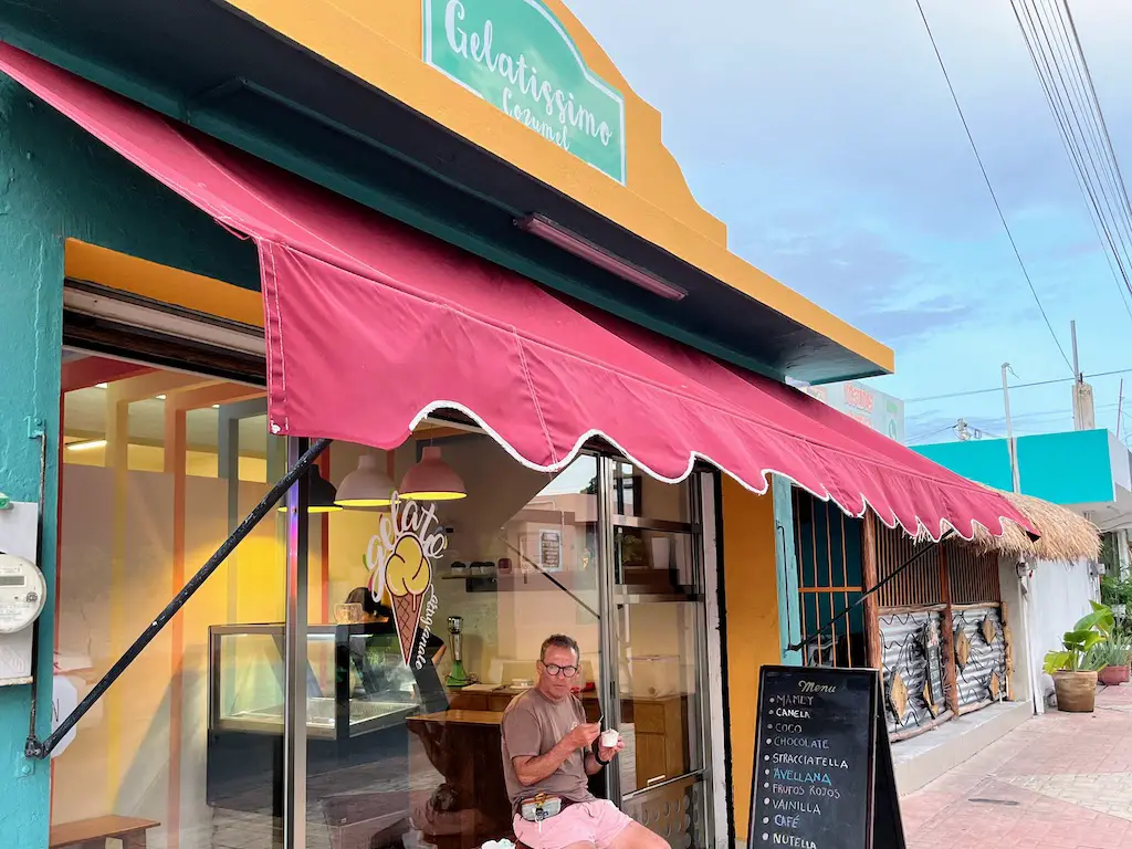 Image we took on our evening ice cream walk in July 2023, shows the facade of the Gelatissimo shop, with awning, list of flavors, and guy enjoying a gelato seated out front. 